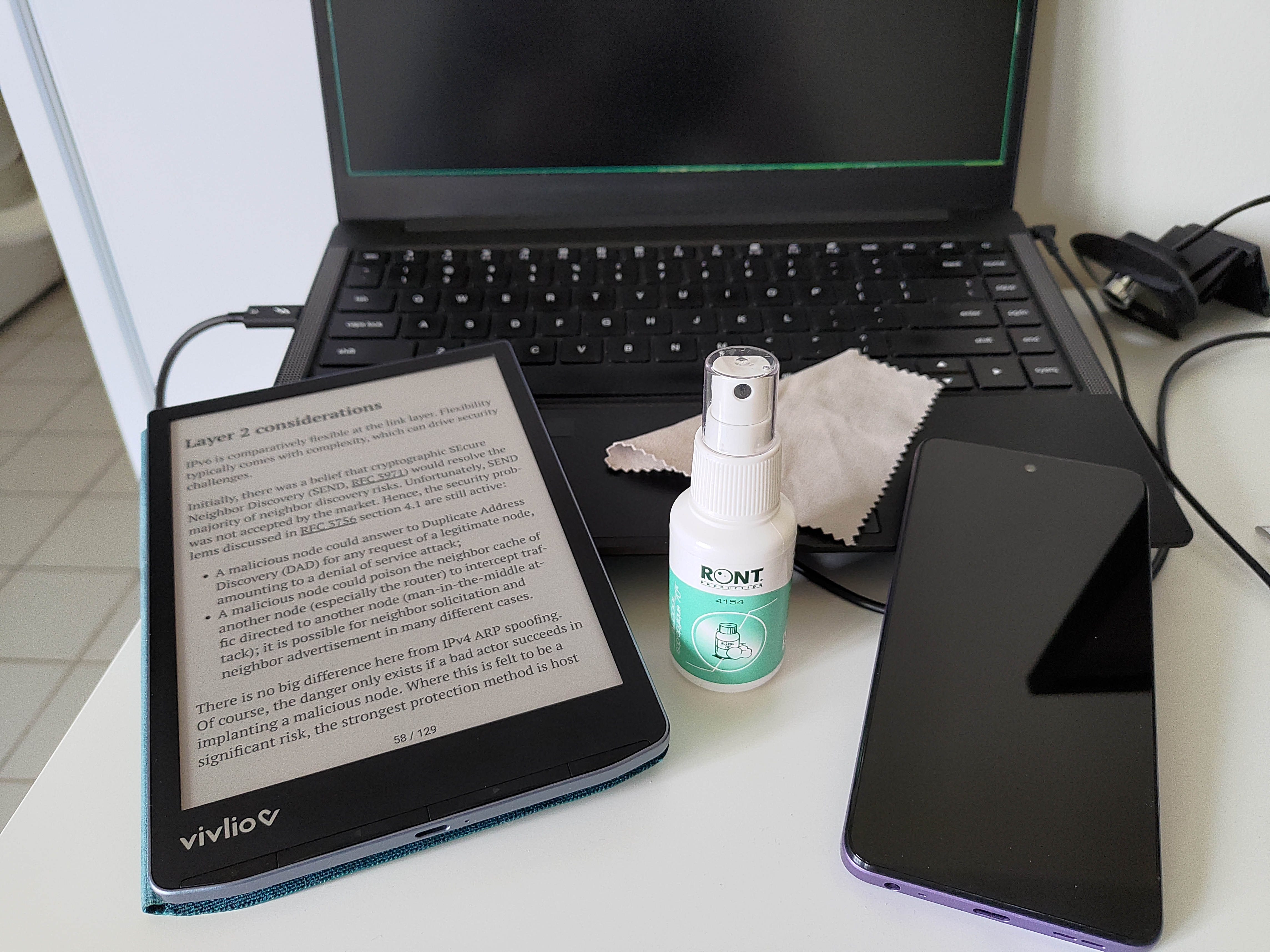 Photographie d'appareils électroniques (ordinateur portable, liseuse, smartphone et webcam) autour d'un pulvérisateur d'alcool isopropylique et d'une lingette. Le tout est posé sur un bureau en mélaminé blanc.