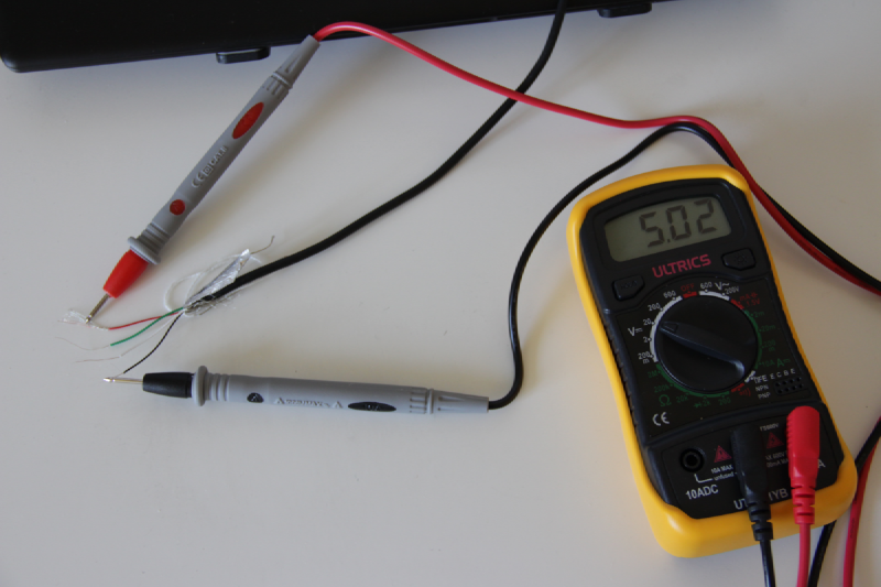 Un câble USB coupé dont sortent 4 fils dénudés : un rouge, un vert, un blanc et un noir. Sur les parties dénudées des fils rouge et noirs sont respectivement posées les bornes rouge et noir d’un multimètre. À côté du câble se trouve un multimètre réglé en mode voltmètre et dont l’écran affiche la valeur 5,02.