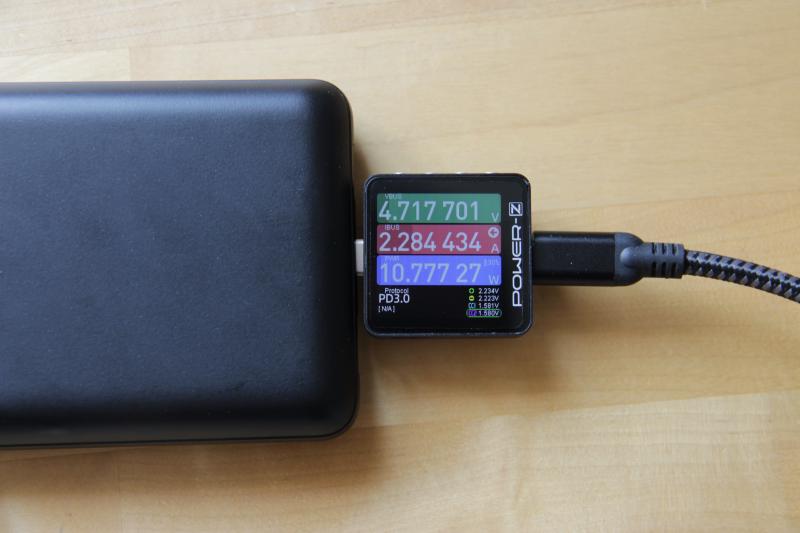 Photographie d'une batterie externe en train d'être chargée à l'aide d'un câble USB-C. Entre la batterie et le câble se trouve un POWER-Z KM003C qui indique une tension de 4,7 V, une intensité de 2,28 A et donc une puissance de 10,7 W. Il est également indiqué l'usage d'USB PD et une flèche indique que le courant va du câble vers la batterie.