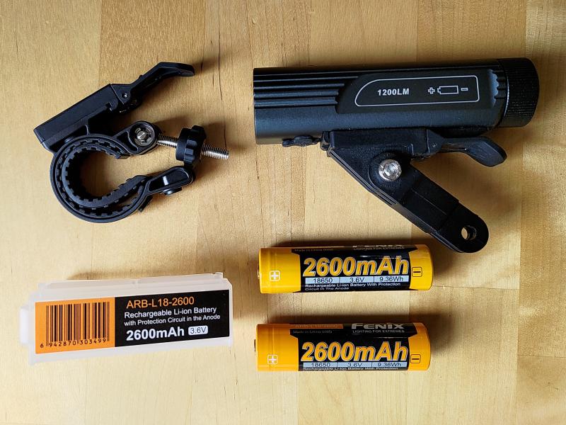 Photographie d'une lampe de vélo BC21R V3.0 posée sur une table en bois. À côté se trouvent un système d'attaque pour guidon ainsi que deux accumulateurs 18650 de 2600 mAh chacun ainsi qu'une boite de rangement en plastique pour un des de ces deux accumulateurs.