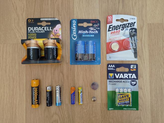 Photographie de piles et accumulateurs de différents formats posés les uns à côté des autres sur deux rangées. Sur la rangée du haut se trouvent, de gauche à droite : un paquet neuf de 2 piles D, un paquet neuf de 2 piles C et un paquet de 2 piles CR2032 dont une seule est présente. Sur la rangée du haut se trouvent, de gauche à droite : un accumulateur 18650, 2 piles AA de marques différentes, 2 piles AAA de marques différentes, une pile CR2032, une pile LR44 et un paquet neuf de 4 accumulateurs AAA.