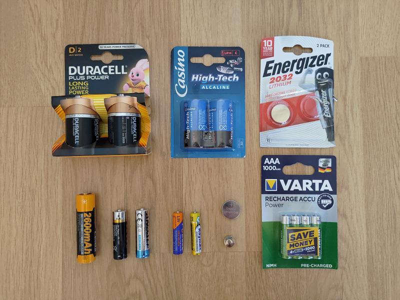 Photographie de piles et accumulateurs de différents formats posés les uns à côté des autres sur deux rangées. Sur la rangée du haut se trouvent, de gauche à droite : un paquet neuf de 2 piles D, un paquet neuf de 2 piles C et un paquet de 2 piles CR2032 dont une seule est présente. Sur la rangée du haut se trouvent, de gauche à droite : un accumulateur 18650, 2 piles AA de marques différentes, 2 piles AAA de marques différentes, une pile CR2032, une pile LR44 et un paquet neuf de 4 accumulateurs AAA.